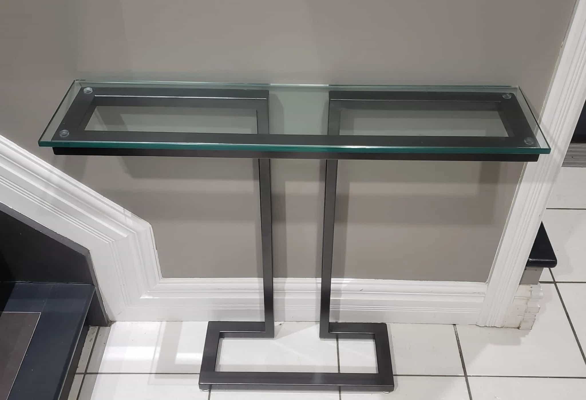 Console table with a clear glass top on two black steel uprights joined by a rectangular floor base