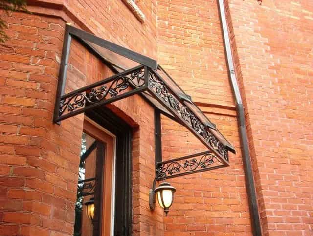 Black wrought iron canopy frame with scrolled side brackets above an entrance door on a red brick wall