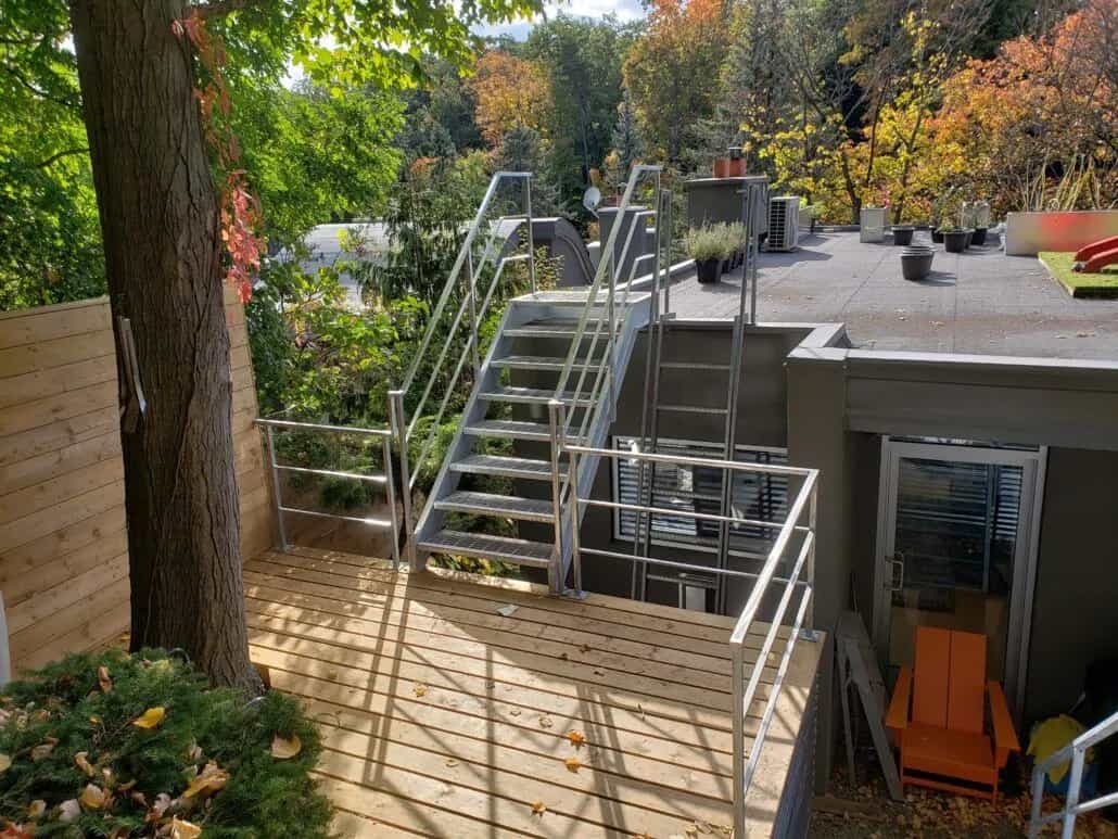 Galvanized steel stair with picket railings climbing from a wood deck to an upper rooftop terrace among trees