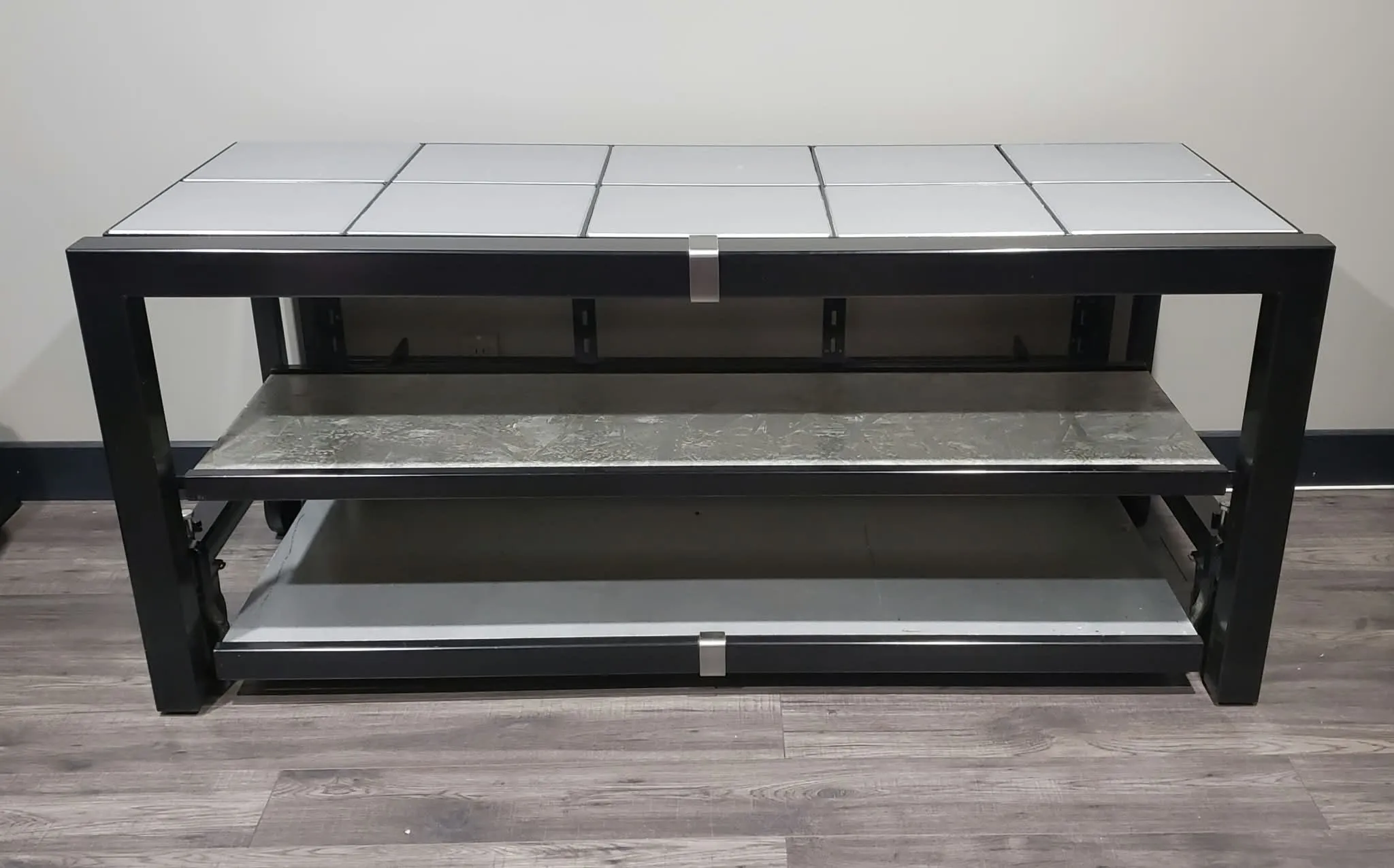Black metal media console with a top made of light grey tiles and two lower shelves, against a wall on a wood-look floor