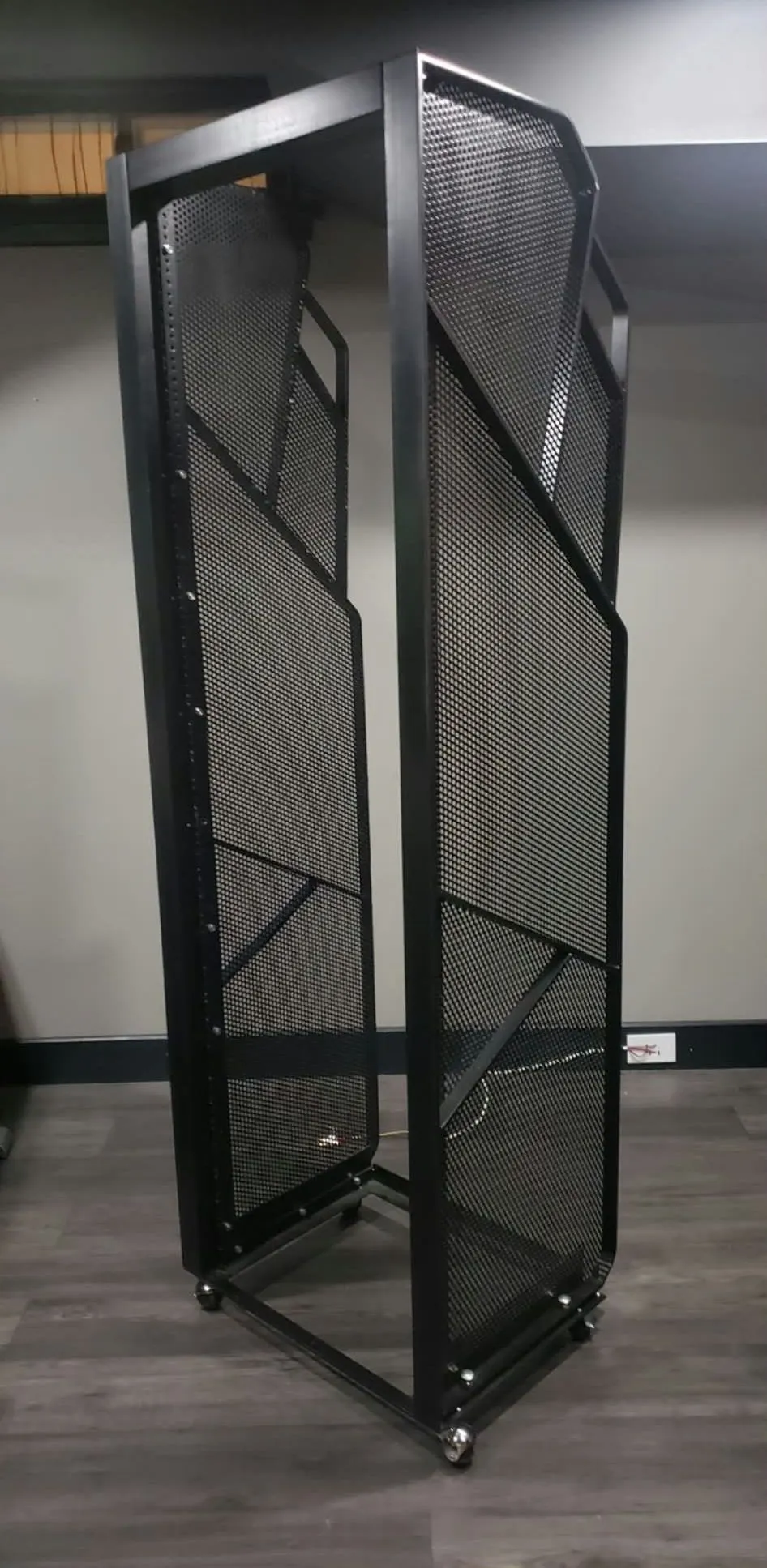 Tall open black metal rack with angled perforated mesh side panels and casters, standing in an empty room