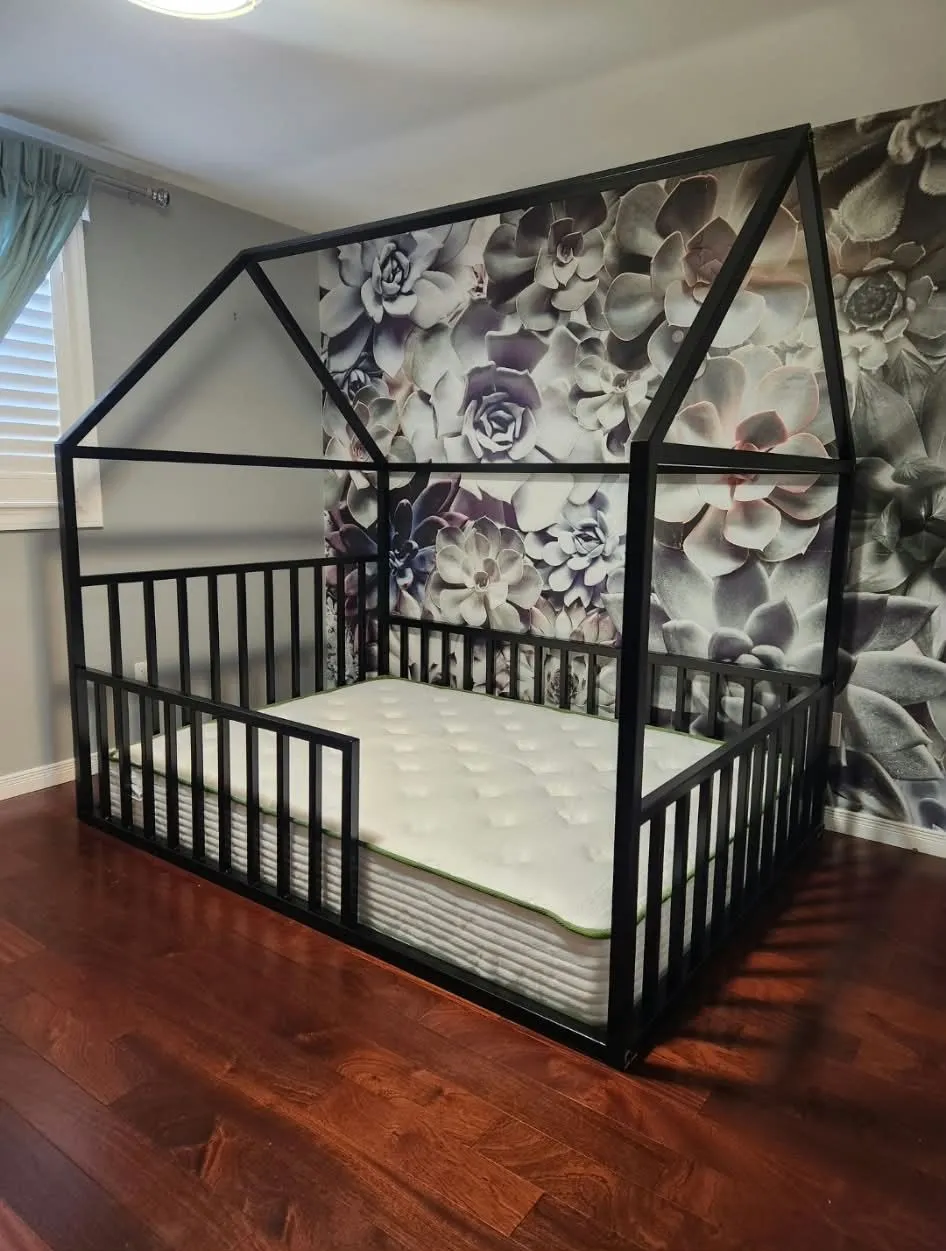 Black metal bed frame shaped like a house outline, with a peaked roof frame and low picket side rails, in a bedroom