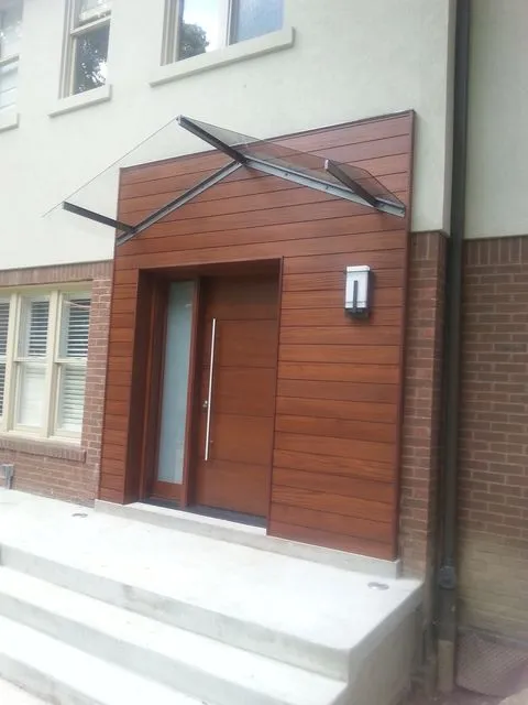 Glass canopy supported by angled steel arms above a wood-clad front door and concrete steps