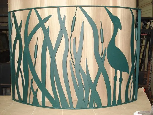 Curved green steel screen cut into tall reeds, cattails, and a heron silhouette