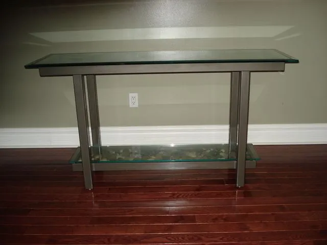 Brushed steel console table with a clear glass top and lower glass shelf against a wall on a hardwood floor