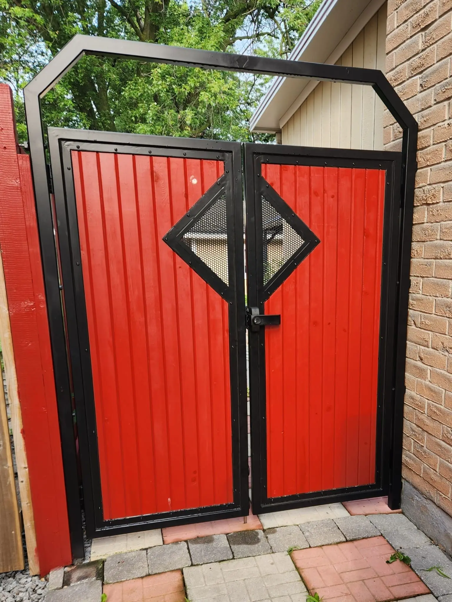 Double gate of red vertical wood boards in a black steel frame with an angled top and a diamond-shaped window