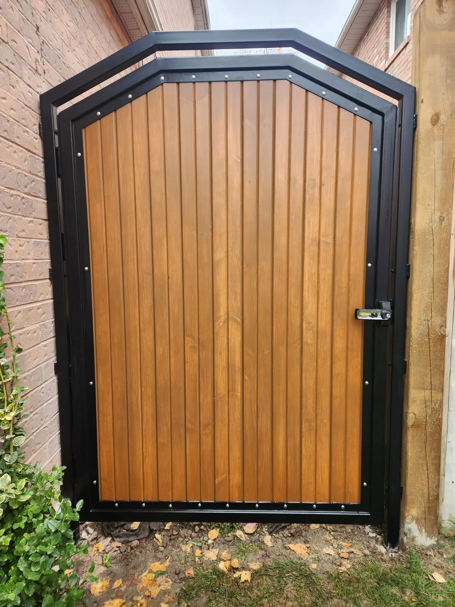 Walk-through gate of vertical cedar boards inside a black steel frame with a peaked top, between brick and a fence