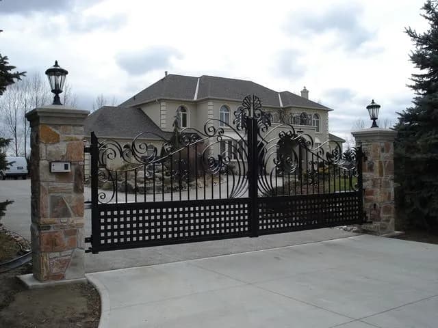 Large black double driveway gate with sweeping scroll tops and lattice lower panels between stone pillars with lanterns