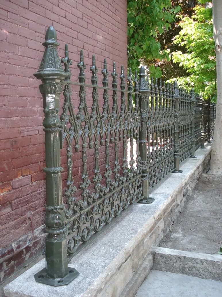 Bronze-green ornamental fence with tall spire posts and pierced scroll panels on a stone wall beside a narrow walkway