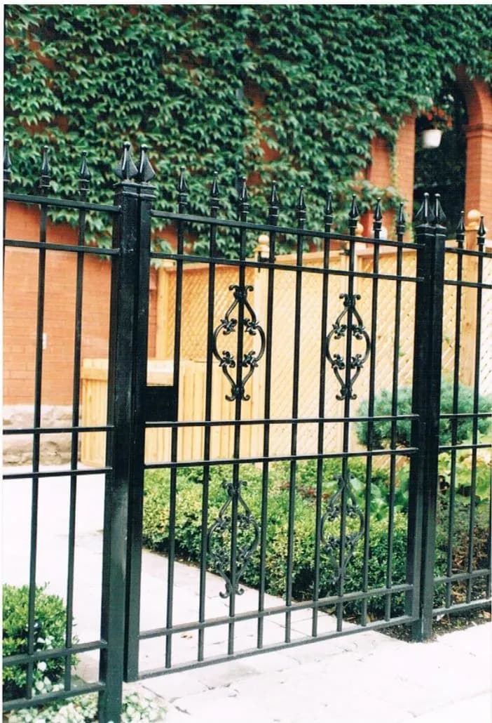 Black wrought iron fence and gate with finial posts and scroll inserts in front of an ivy-covered brick wall