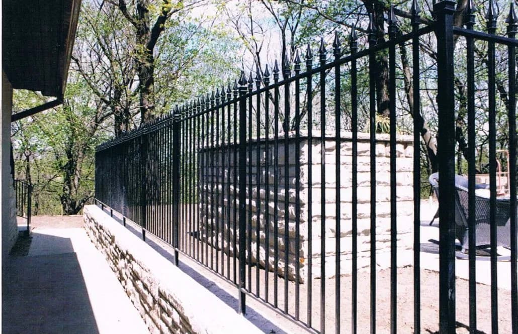 Tall black spear-top picket fence running along the top of a stone retaining wall beside a paved walkway