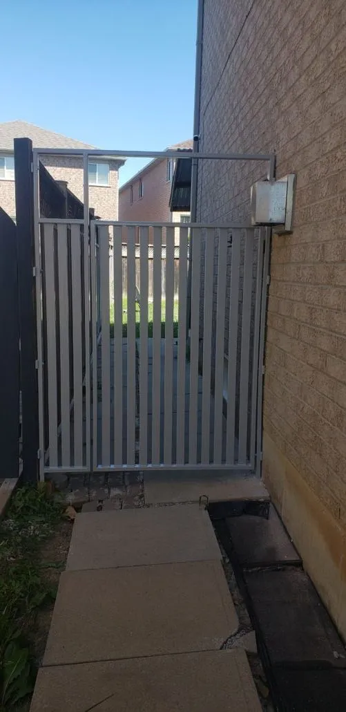 Grey aluminum walk-through gate with vertical pickets in a narrow side yard between a brick house and its neighbour