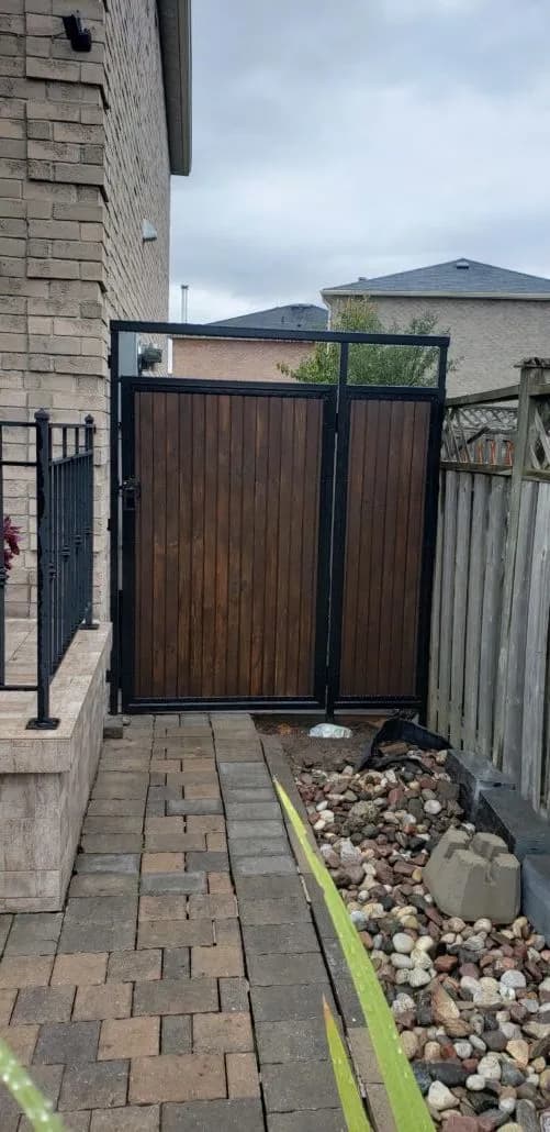 Black steel gate frame with stained wood panels across an interlock side path, next to a black railing and river stones