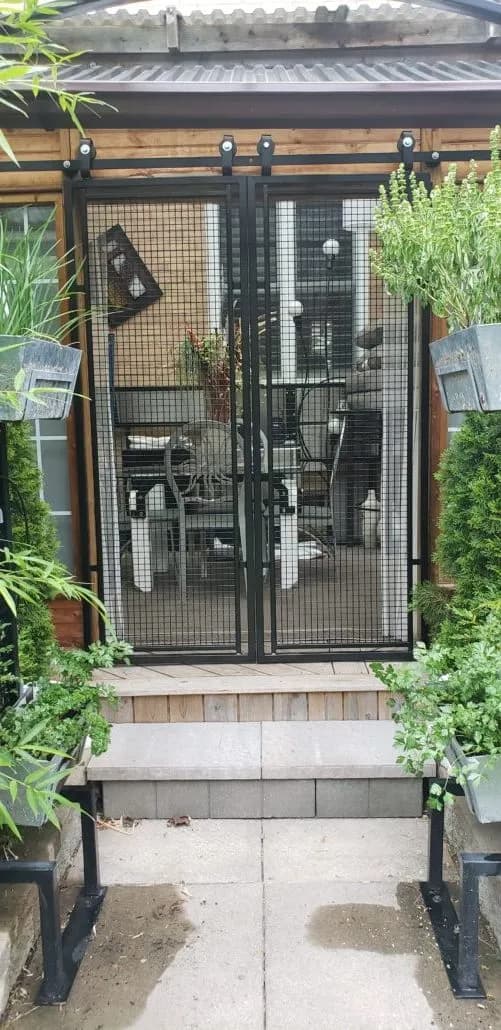Black steel double doors with wire mesh panels framing the entrance to a patio, with potted plants on either side