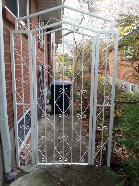 White-painted iron gate and arbour with an arched top and scroll details over a side path beside a brick house
