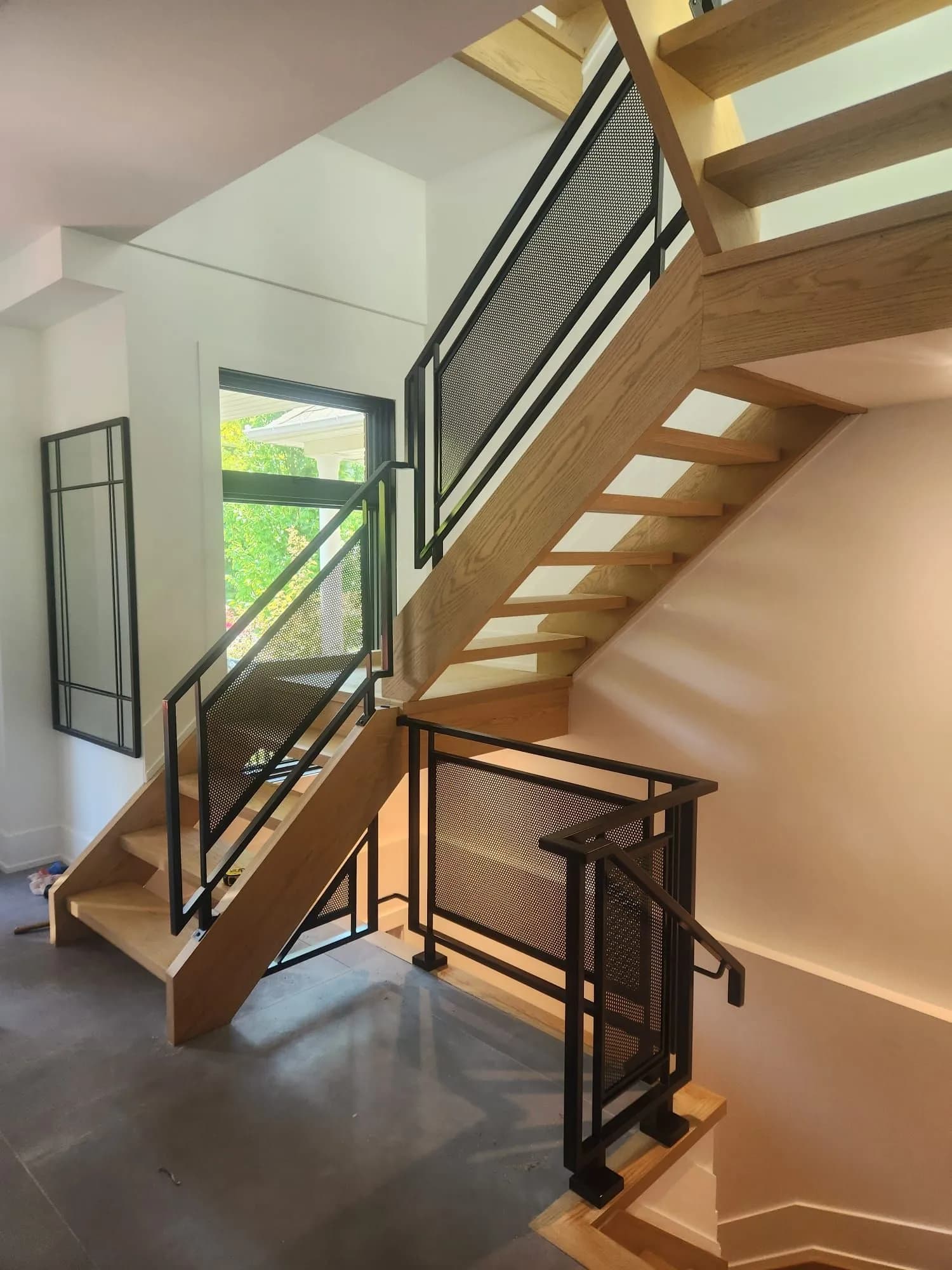 Open-riser oak staircase with a black steel railing holding perforated metal panels, beside a tall black-framed window
