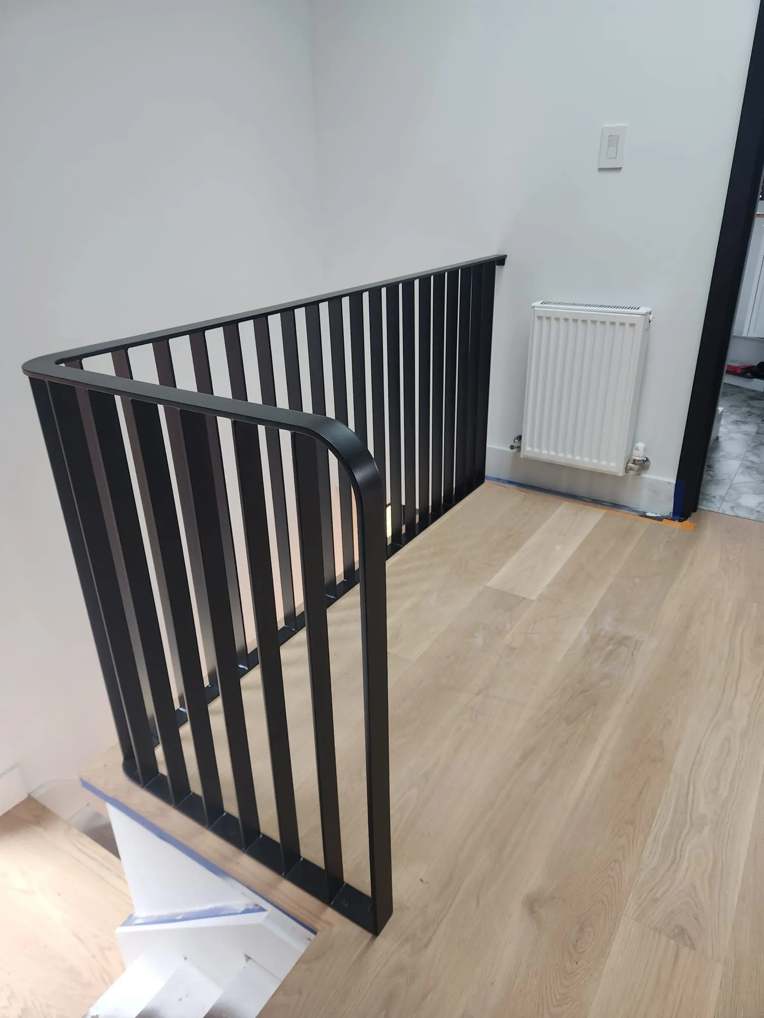 Black steel guard with slim vertical bars and a smoothly bent corner rail along an upper landing with light oak floors