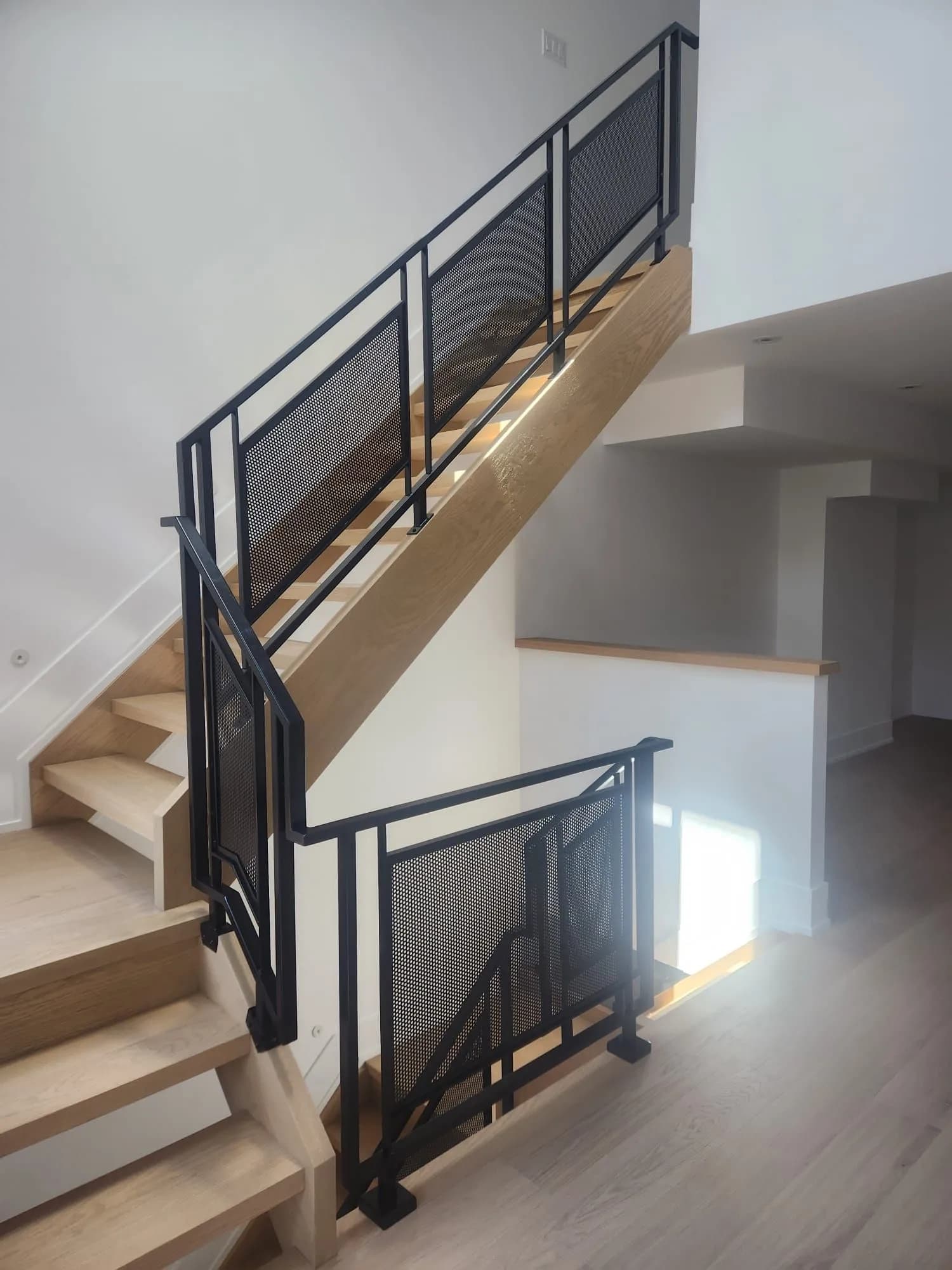 Black steel frame railing with perforated metal infill following an oak staircase up to a landing in a bright new home