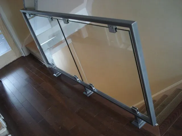 Clear glass panels in a brushed stainless steel frame with base clamps, guarding an upper landing over dark hardwood