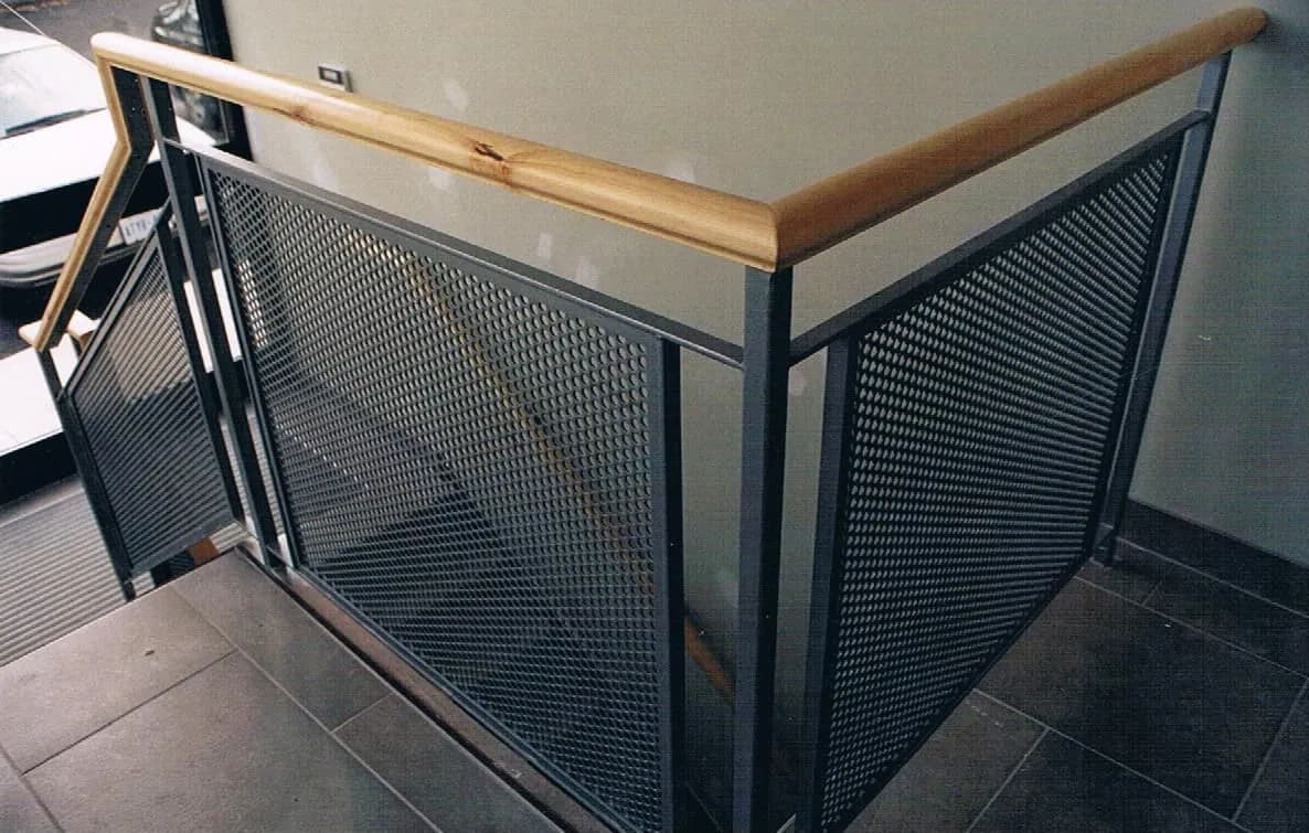 Dark steel frame with perforated metal infill and a light wood handrail wrapping a stair opening on a tiled floor
