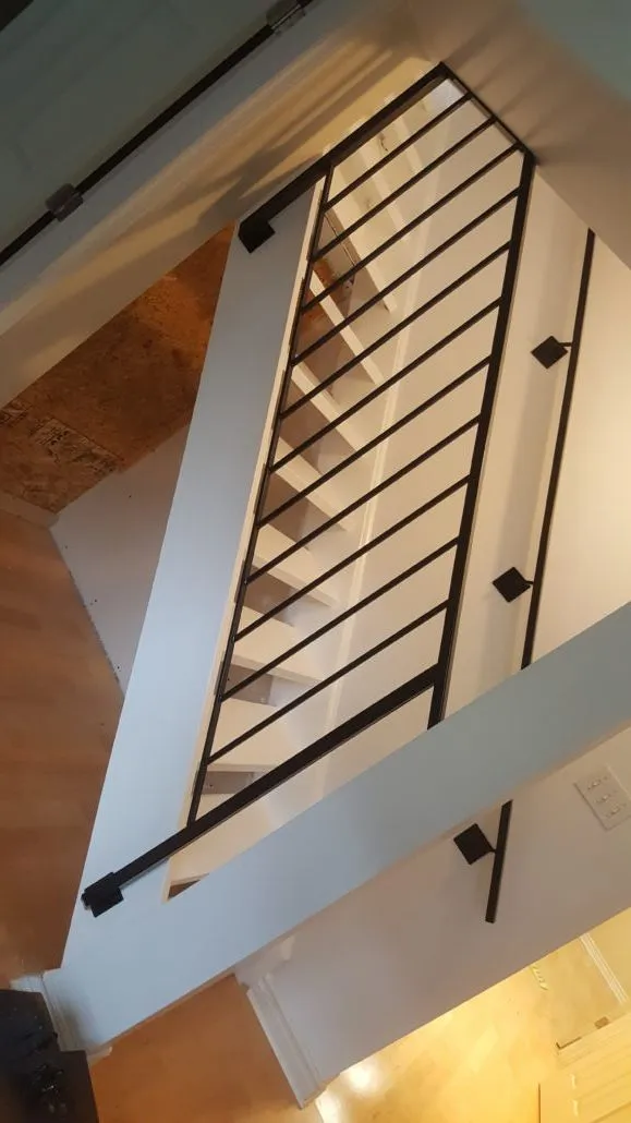 Black steel stair railing with evenly spaced vertical bars and a wall-mounted handrail, looking down a stairwell