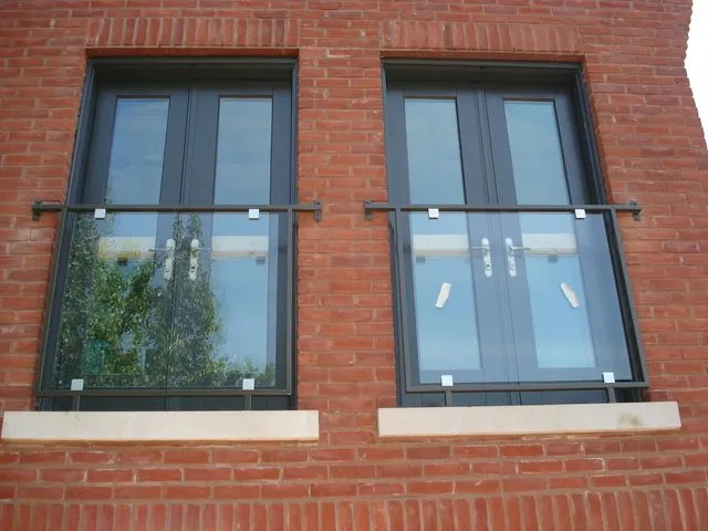 Two frameless glass Juliet balconies with slim black top rails across tall French doors on a red brick wall