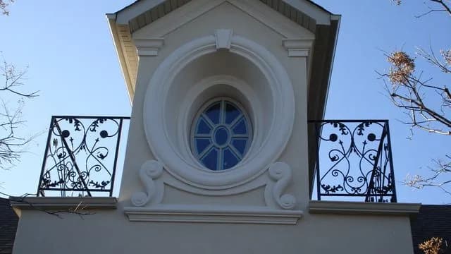 Two black wrought iron scroll Juliet railings on either side of a round window in a white stucco dormer