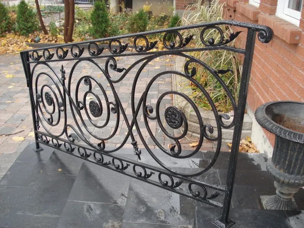 Black wrought iron step railing filled with large spiral scrolls and rosettes on grey stone steps beside a stone urn
