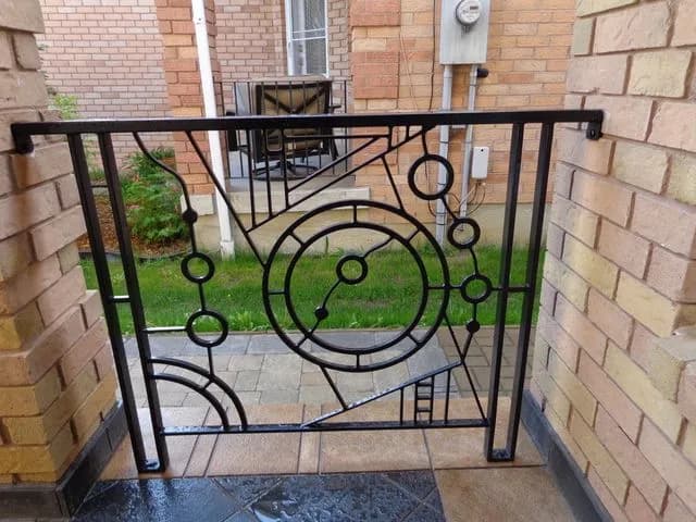 Black steel entry railing with concentric circles and angled lines in an Art Deco pattern between two brick walls