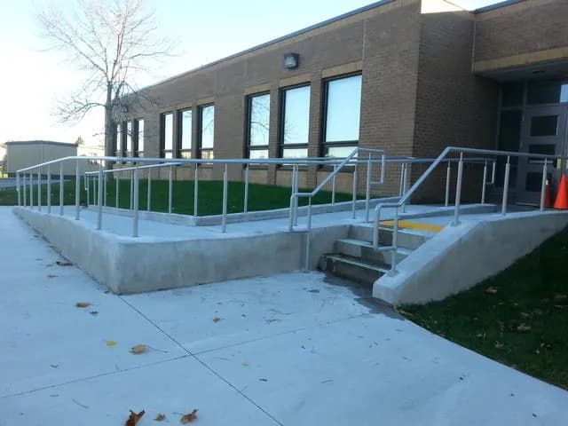 Galvanized steel handrails along a long concrete ramp and short steps leading to a brick commercial building
