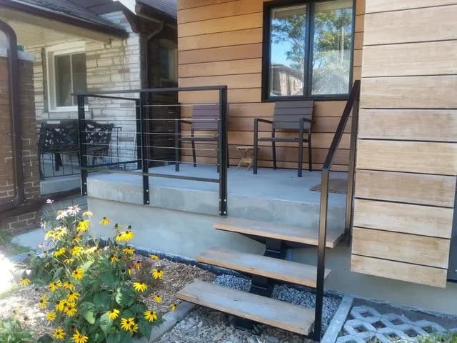 Black steel porch railing with thin horizontal rods above floating wood steps and a flower bed