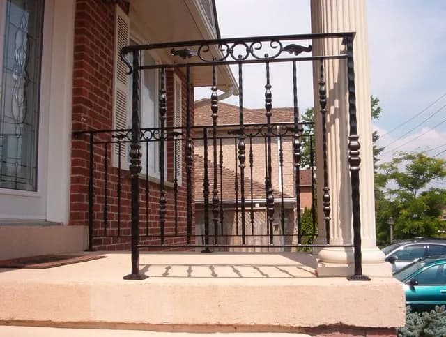 Black iron porch railing with turned decorative balusters and a scroll top panel between a white column and brick