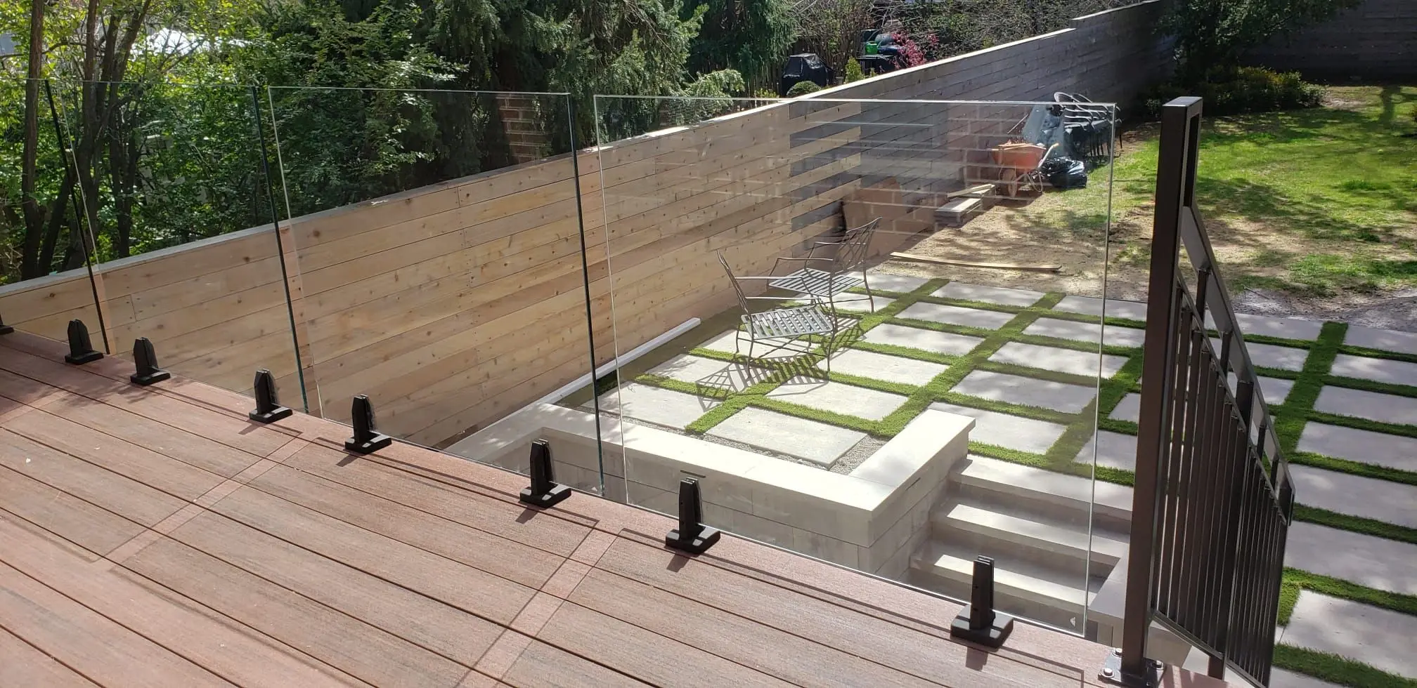 Clear glass panels held by black spigot clamps along the edge of a composite deck overlooking a backyard patio