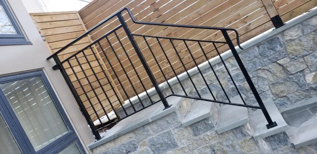 Black steel railing with vertical bars and a flat top rail along stone-clad steps next to a horizontal cedar fence