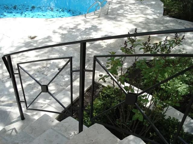 Black steel railing with X-braced square panels beside concrete steps leading down to a backyard pool