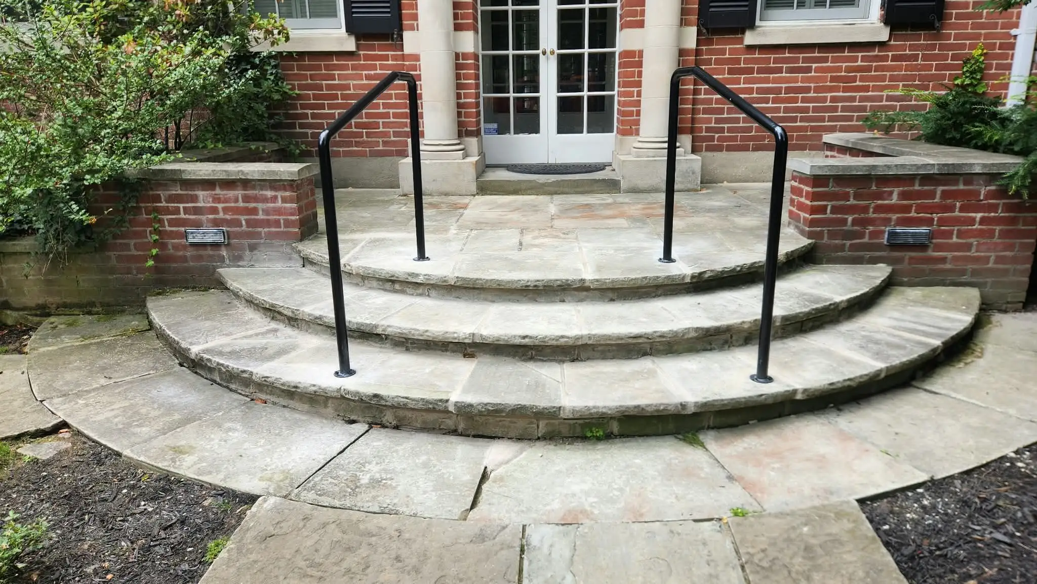 Two black metal handrails on a wide set of curved natural stone steps leading up to French doors between brick walls
