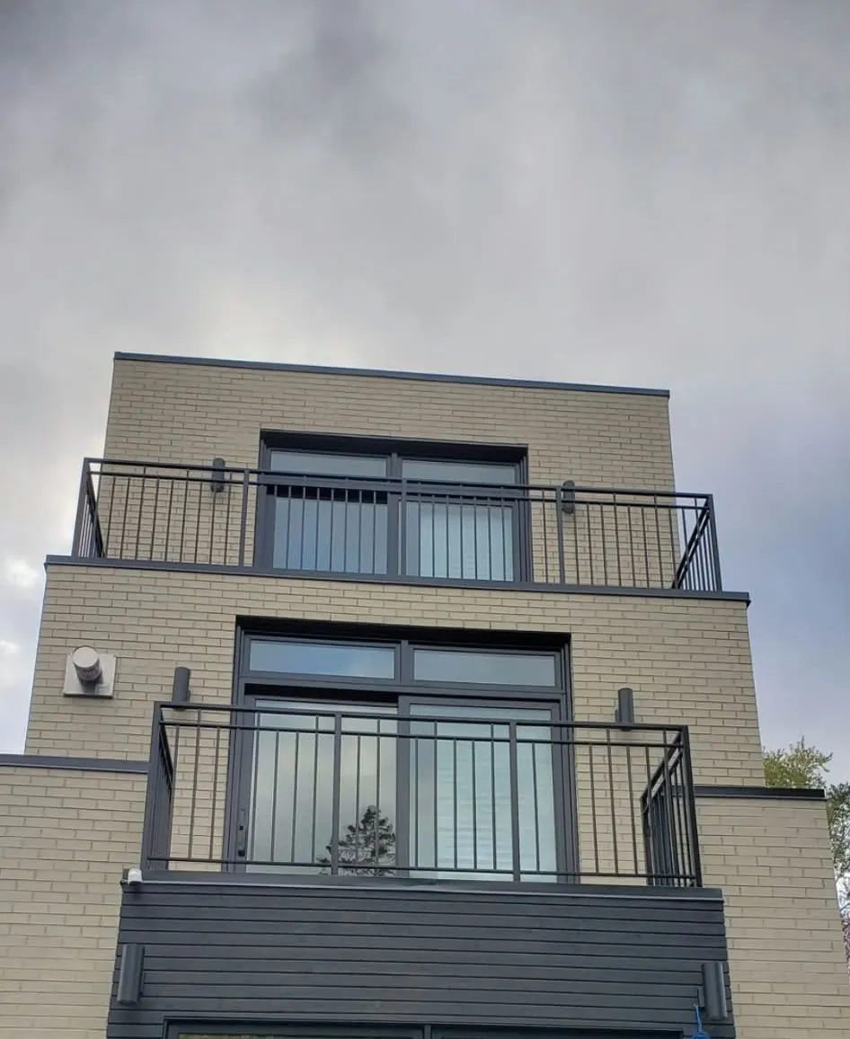 Two black metal picket balcony railings across full-height windows on the upper floors of a modern brick and dark-clad house