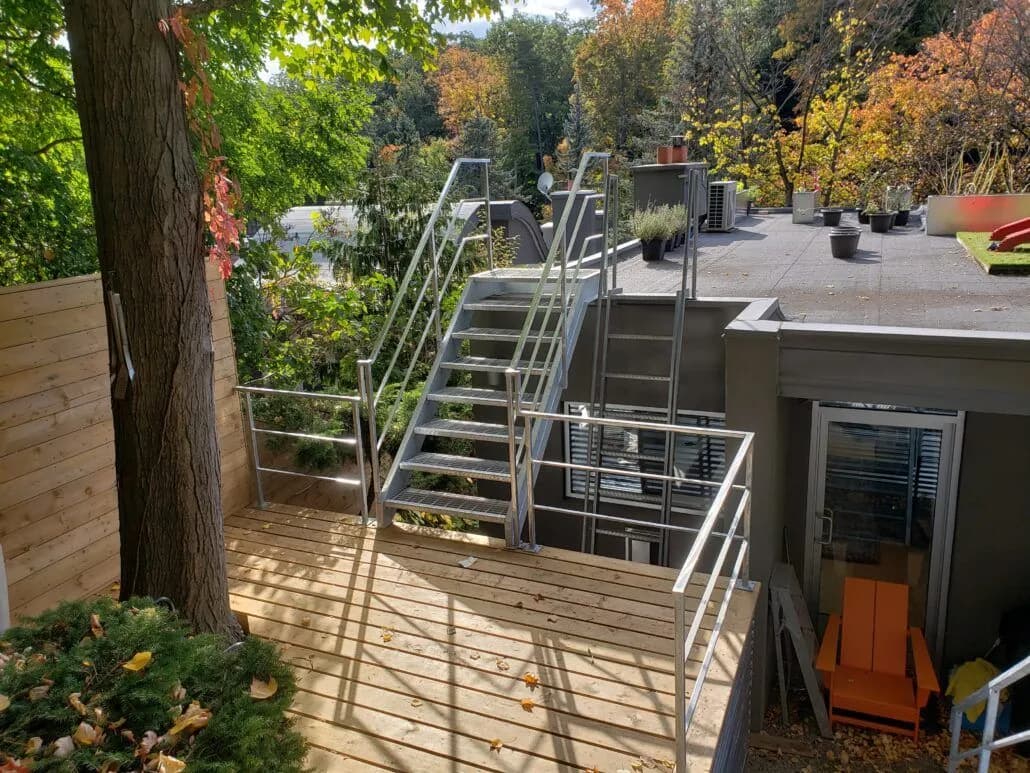 Galvanized steel stair with picket railings climbing from a wood deck to an upper rooftop terrace among trees