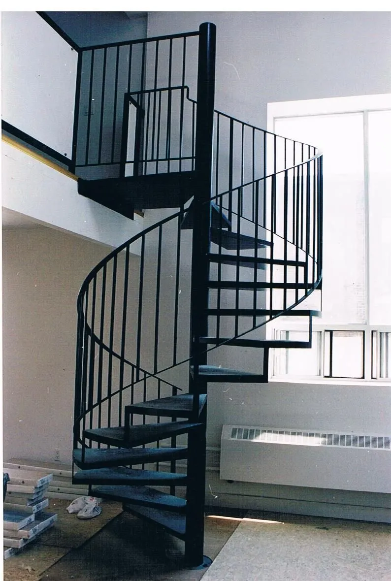 Black steel spiral staircase with open treads and a vertical picket railing rising to a loft guard in a bright room