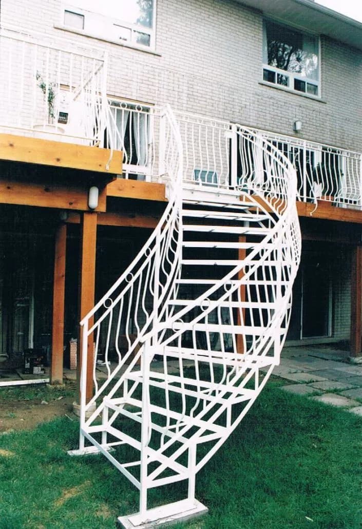 White-painted steel exterior staircase curving down from a wood deck to the lawn, with bowed ornamental railings